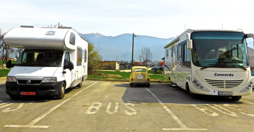A small yellow car is shown in a huge parking space. Either side of it are two huge white buses.