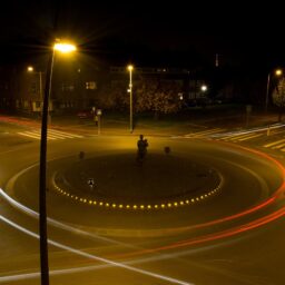 A long-exposure photograph of a roundabout at night. No cars can be seen, but the trail left by their white and red headlights and taillights remains.