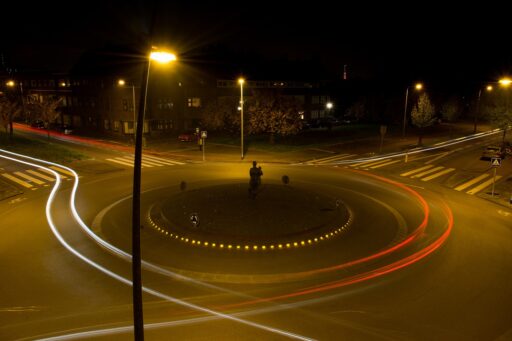 A long-exposure photograph of a roundabout at night. No cars can be seen, but the trail left by their white and red headlights and taillights remains.