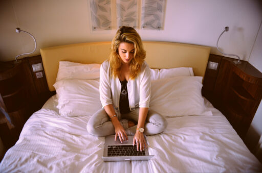 A professional woman working on a laptop while sitting cross-legged on a bed in a hotel room.